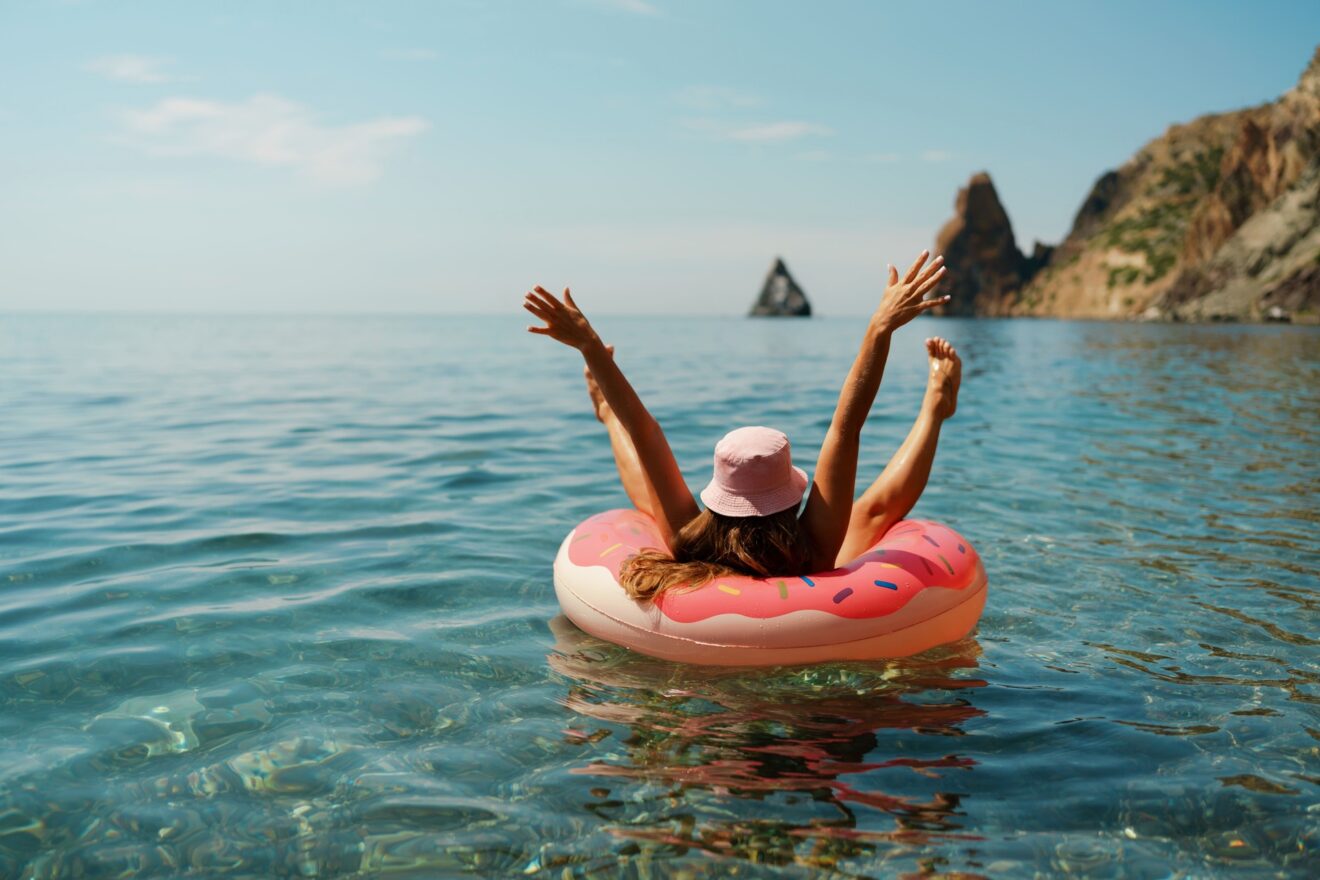 Woman relaxing on a pink donut-shaped float in crystal-clear water, enjoying a sunny day by the rocky coastline.