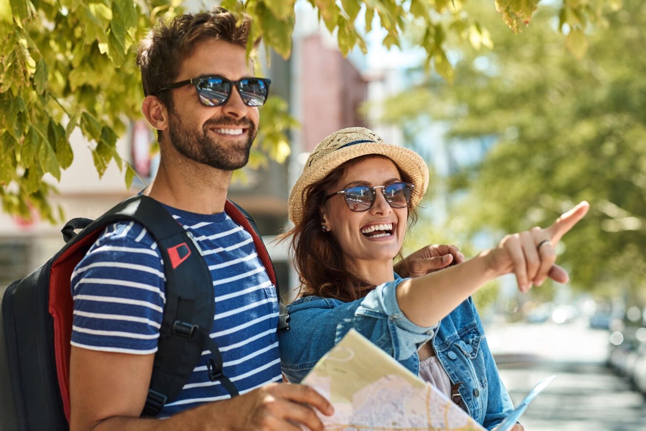 A smiling couple of travelers enjoying a sunny day outdoors, holding a map and pointing towards a destination. Perfect image to represent adventurous tourism.